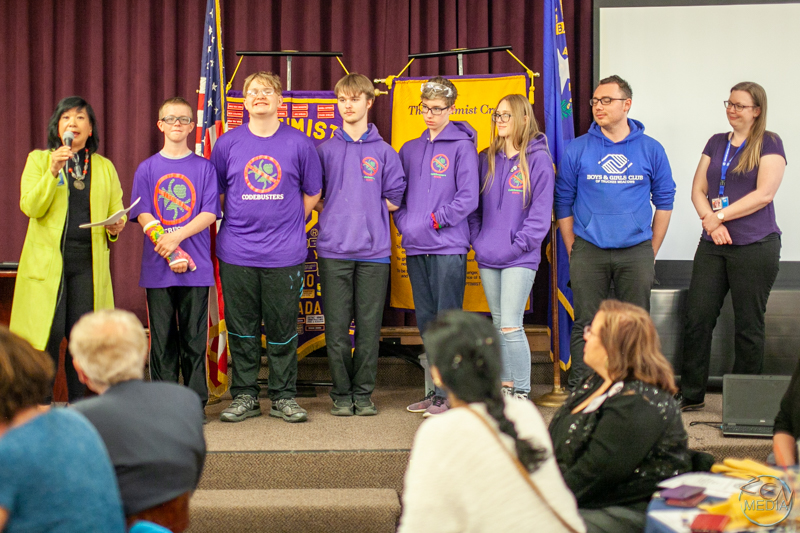 A team of five junior robotic builders, four males and one female, each wearing matching purple tops with a team logo, and their adult male captain on stage, as they receive an award.