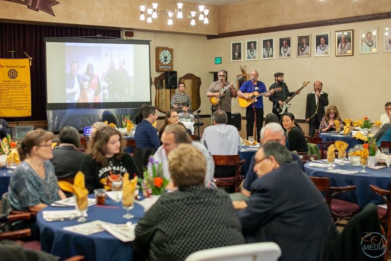 Members and participants of various Optimist Club of Reno programs, seated at banquest tables, talking and listening to music from a band on a stage.